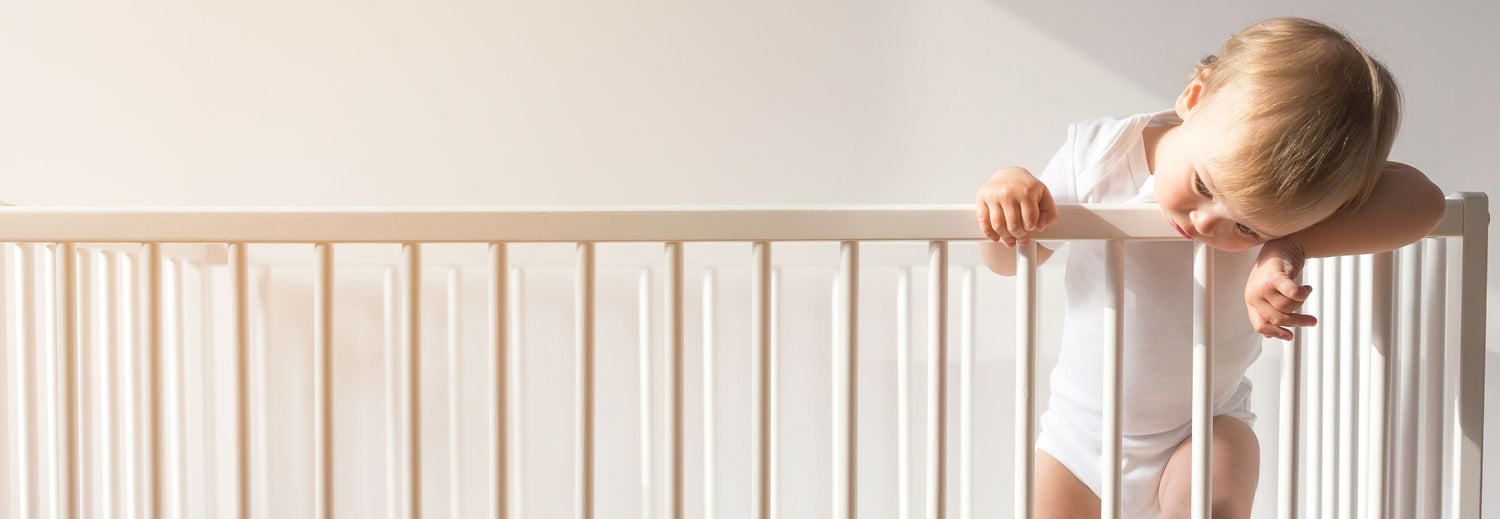 Baby standing in a white crib, leaning over the rail in a minimalist, light-filled nursery