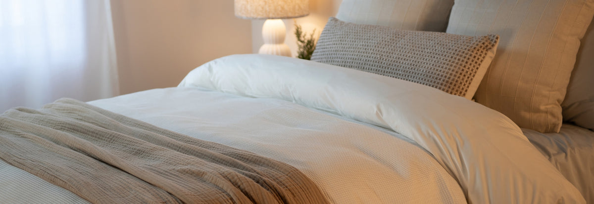 A neatly made bed with white bedding and a textured beige throw blanket, styled with neutral pillows in a softly lit bedroom setting.