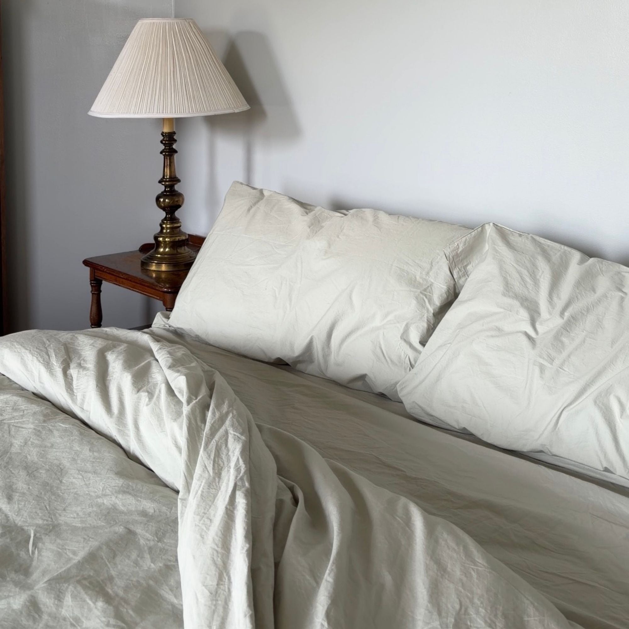 Neatly made bed with white bedding and pillows, nightstand with lamp in the corner.