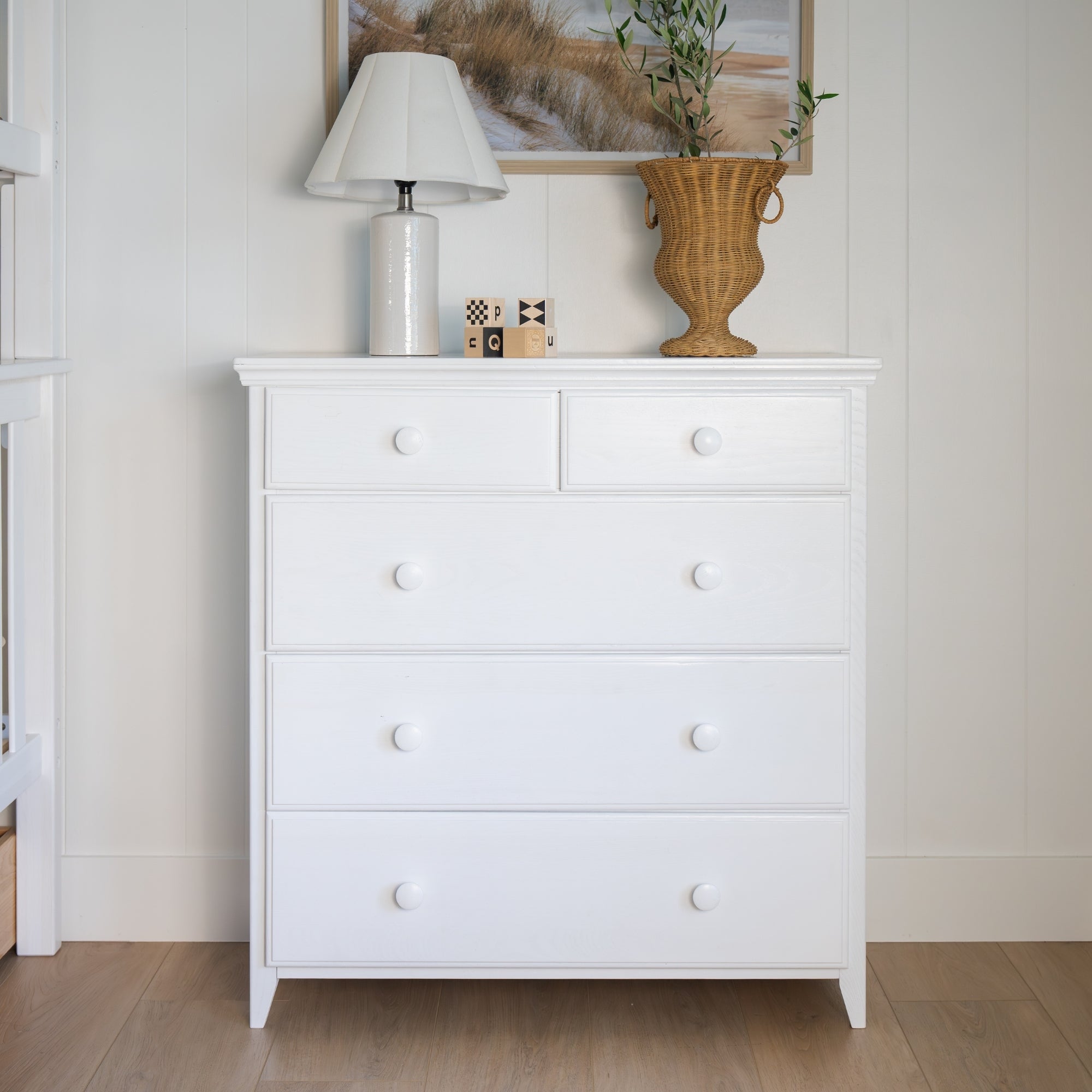 A white wooden five-drawer dresser with round white knobs, styled with a lamp, small decorative blocks, and a wicker vase #color_washed white