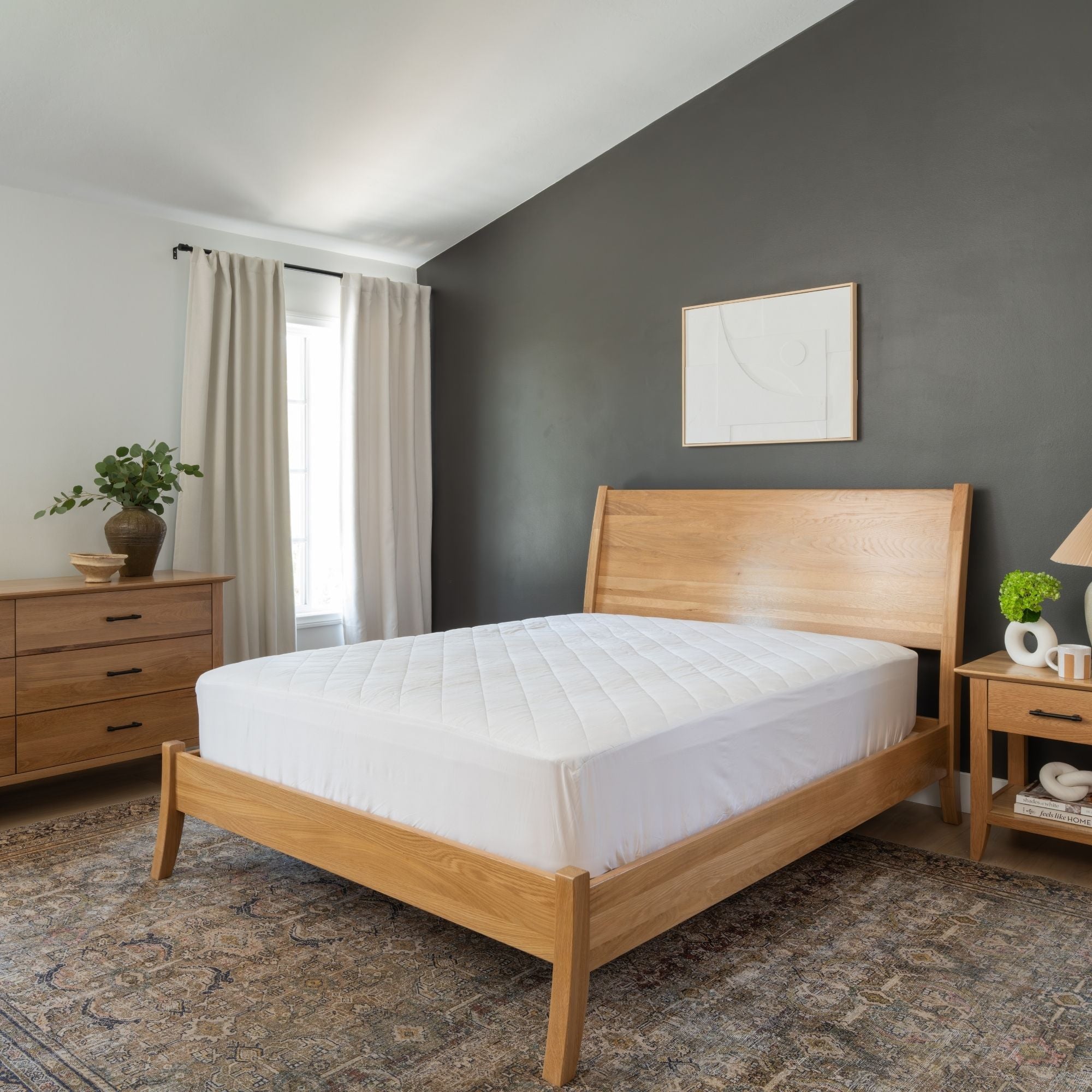A light oak wood bed frame with a white quilted mattress pad, styled in a modern bedroom with a dark gray accent wall, beige curtains, and a patterned rug.
