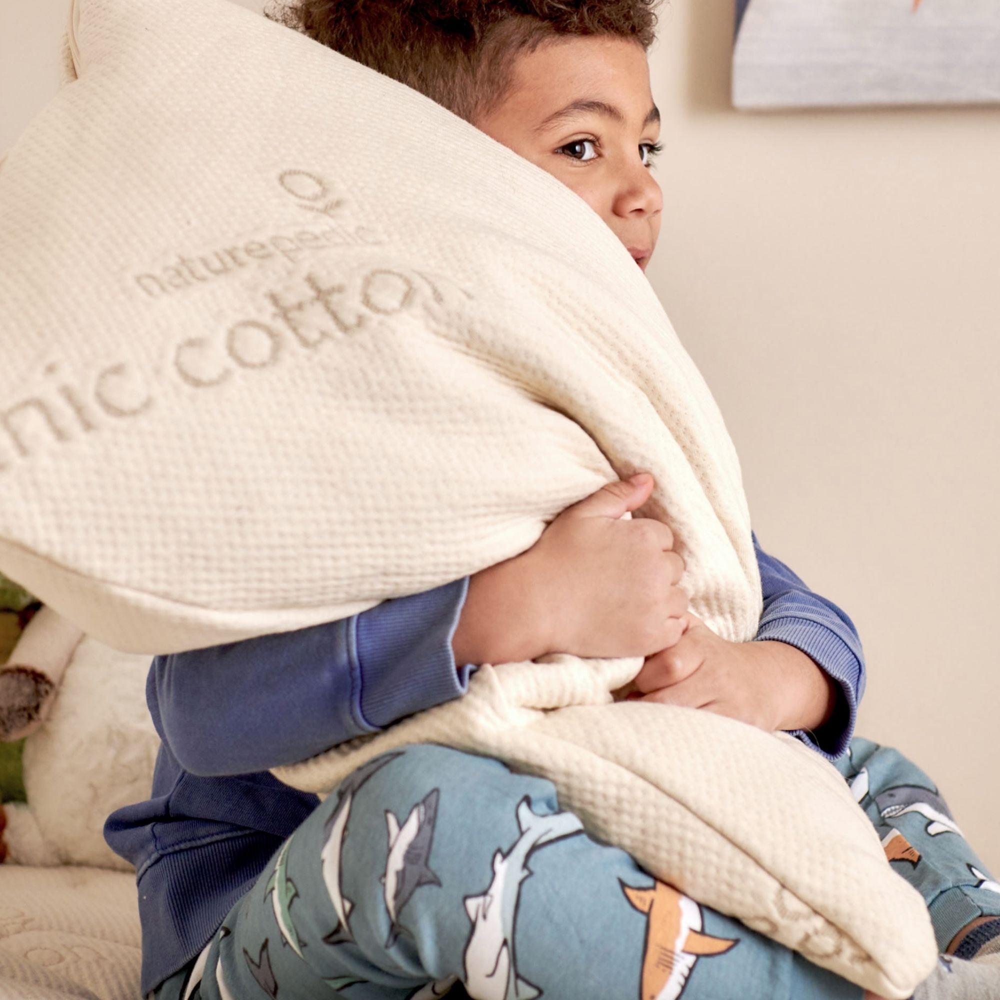 Child holding a pillow with 'naturepedic organic cotton' text, sitting on a mattress.