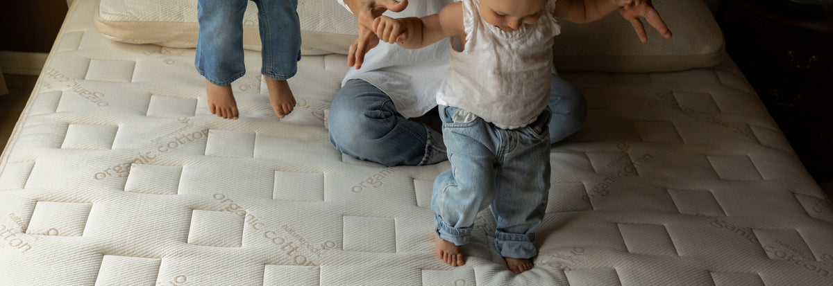 A toddler in a white t-shirt and blue jeans stands on a bare mattress while an adult in a white shirt and blue jeans holds their hand. Another person's legs are visible jumping on the mattress in the background.